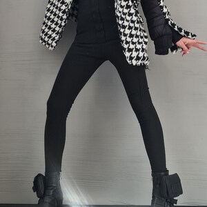 May include: A black turtleneck sweater, black leggings, and a black and white houndstooth jacket with quilted black sleeves. The outfit is paired with black leather ankle boots featuring side buckles, thick lug soles, and small attached pouches.