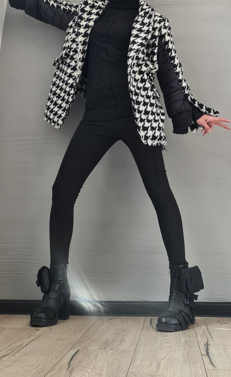 May include: A black turtleneck sweater, black leggings, and a black and white houndstooth jacket with quilted black sleeves. The outfit is paired with black leather ankle boots featuring side buckles, thick lug soles, and small attached pouches.