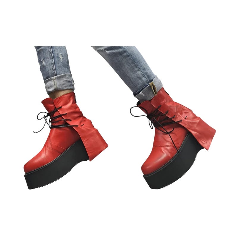 K&ouml;nnte beinhalten: Rote Leder-Plateau-Stiefel mit schwarzen Schn&uuml;rsenkeln und einer dicken schwarzen Sohle. Die Stiefel haben ein einzigartiges Design mit einer Klappe, die sich an der Seite erstreckt. Die Stiefel werden mit einer blauen Jeans getragen.