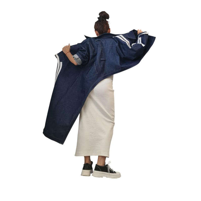 May include: A long, dark blue denim jacket with white stripes on the sleeves and back. The jacket is worn over a cream-colored dress. The person is wearing black and white sneakers.