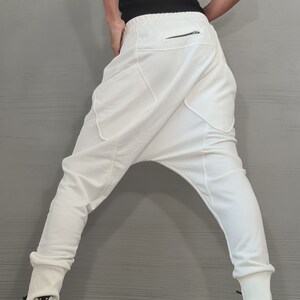 Op de afbeelding: Witte harembroek met een lage kruis en een ritszak. De broek wordt gecombineerd met een zwarte tanktop met witte bies en witte en zwarte sneakers.