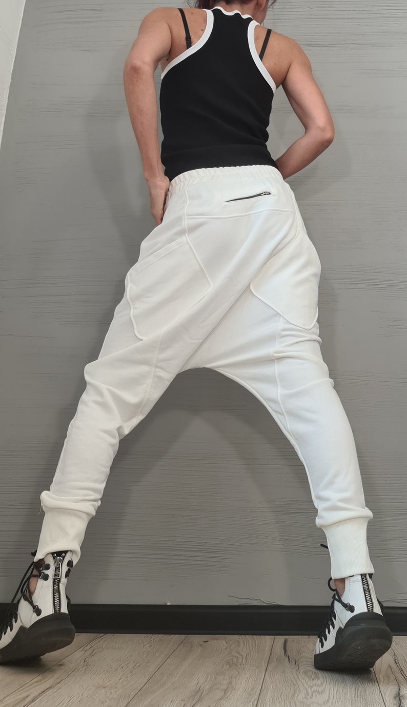 Op de afbeelding: Witte harembroek met een lage kruis en een ritszak. De broek wordt gecombineerd met een zwarte tanktop met witte bies en witte en zwarte sneakers.