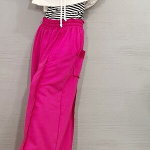 May include: A woman's outfit featuring a white hooded sweatshirt, a black and white striped long-sleeve shirt, and bright pink sweatpants. The sweatpants have a pocket on the side and a drawstring waist. The outfit is casual and comfortable.
