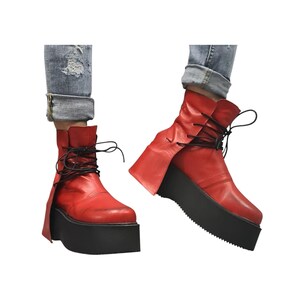 K&ouml;nnte beinhalten: Rote Stiefeletten mit schwarzen Plateausohlen und schwarzen Schn&uuml;rsenkeln. Die Stiefel haben ein einzigartiges Design mit einer umgeklappten Klappe an der Seite. Die Plateausohlen sind dick und verleihen H&ouml;he. Die Stiefel werden mit blauen Jeans getragen.