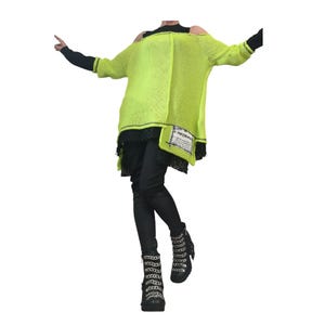 May include: A neon yellow open-knit sweater with cold-shoulder cutouts, layered over a black long-sleeve top and black pants. The sweater features a white rectangular patch with black text on the lower front and black lace trim at the hem.