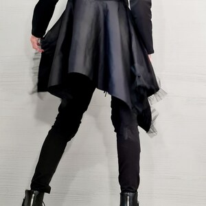 May include: Black long-sleeved top with a flared, asymmetrical skirt. The skirt has a satin-like appearance and is trimmed with a delicate, sheer fabric. The outfit is paired with black pants and black ankle boots with zippers.