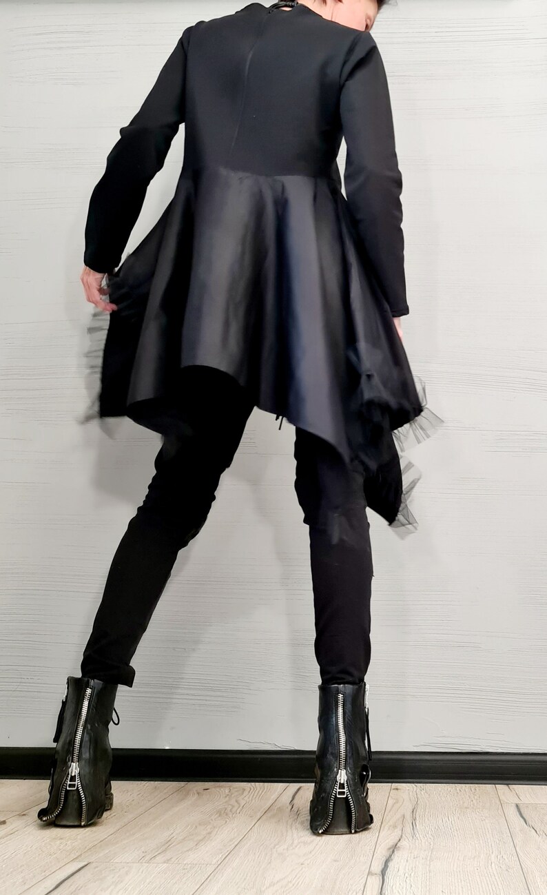 May include: Black long-sleeved top with a flared, asymmetrical skirt. The skirt has a satin-like appearance and is trimmed with a delicate, sheer fabric. The outfit is paired with black pants and black ankle boots with zippers.
