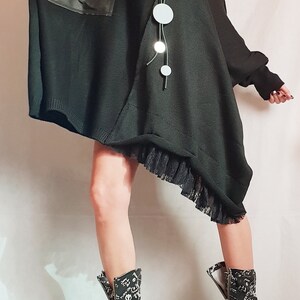 May include: An oversized black knit tunic featuring a smooth black pocket, a black lace hem, and a long necklace with circular silver pendants. The outfit is paired with black mid-calf boots covered in white graffiti-style text and symbols.