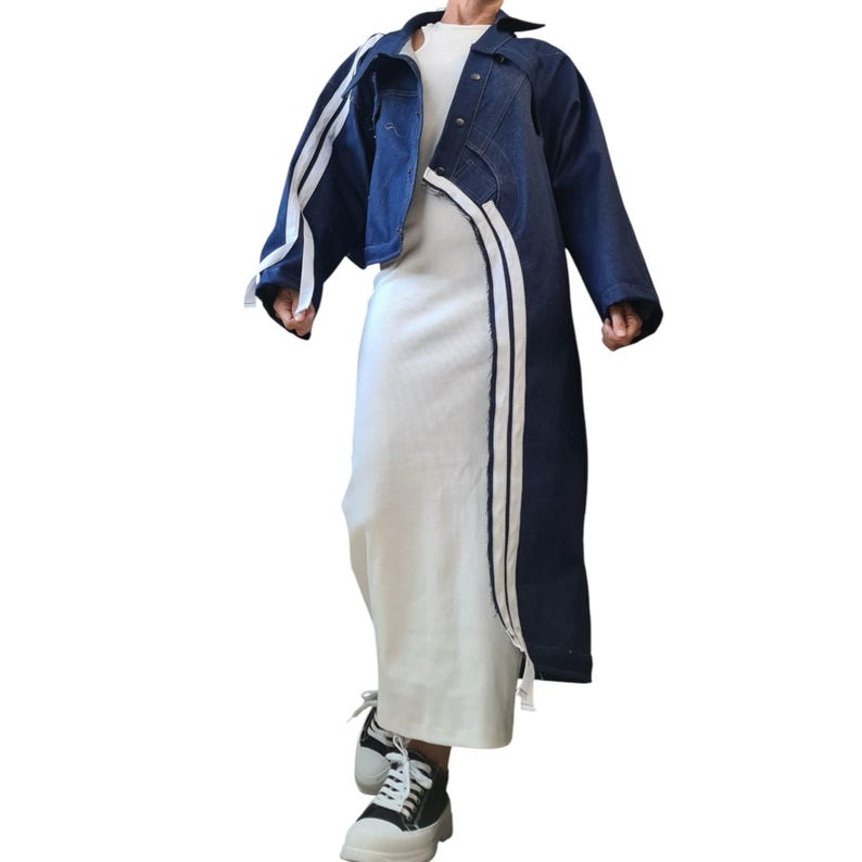 May include: A dark blue denim jacket with white stripes down the sleeves and front. The jacket is worn over a white dress. The outfit is completed with black and white sneakers.