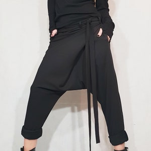 May include: Black ensemble featuring a long-sleeved top with shoulder cutouts and a high neckline. The outfit includes draped, loose-fitting pants with a waist tie and rolled cuffs. Black ankle boots with buckles and laces complete the look.