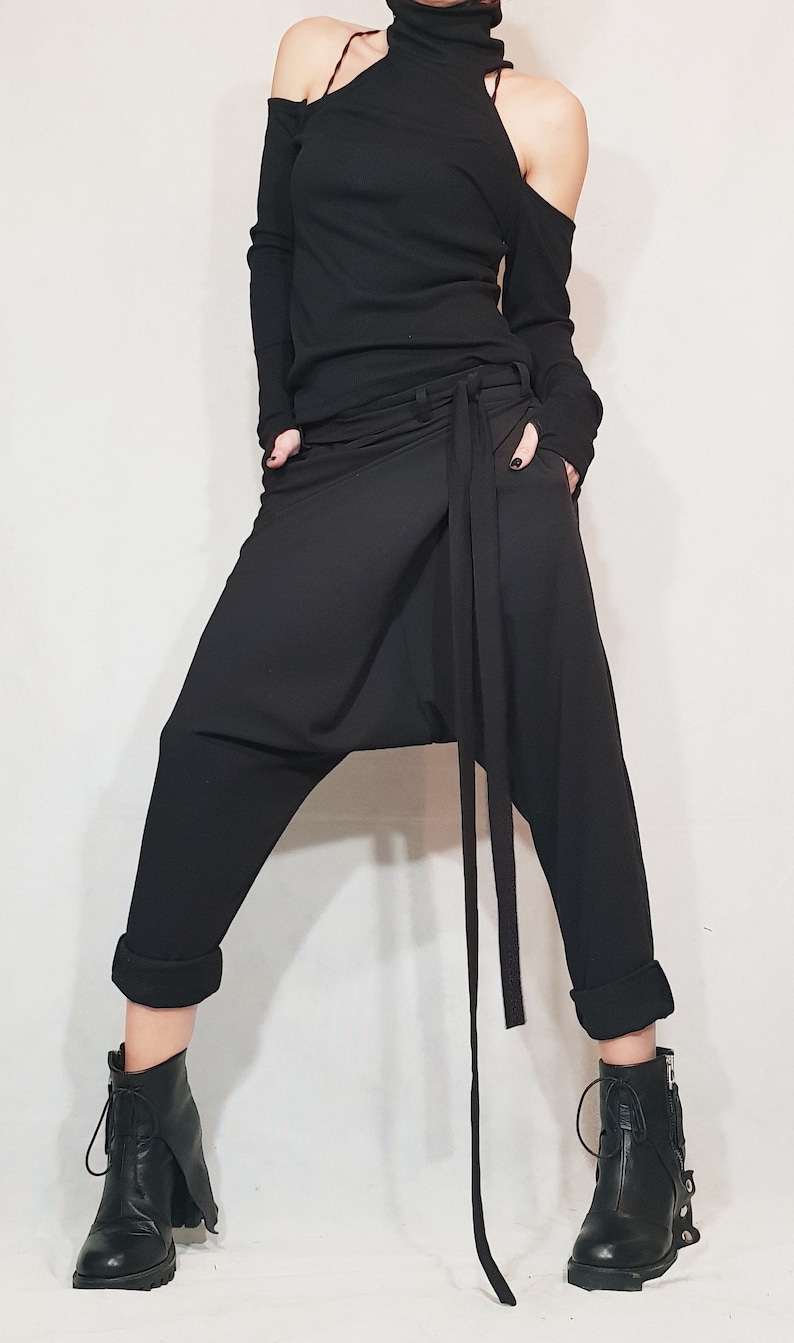 May include: Black ensemble featuring a long-sleeved top with shoulder cutouts and a high neckline. The outfit includes draped, loose-fitting pants with a waist tie and rolled cuffs. Black ankle boots with buckles and laces complete the look.