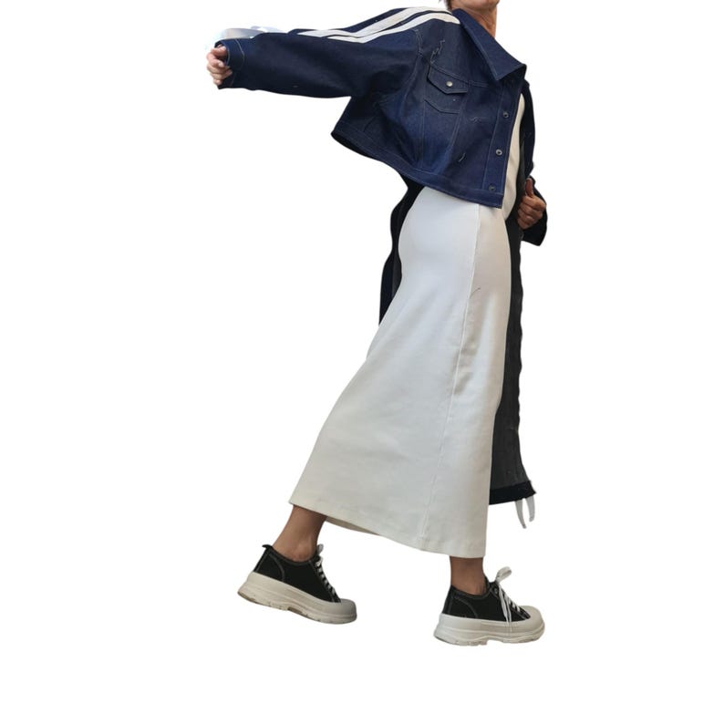 May include: A dark blue denim jacket with white stripes on the sleeves, worn over a white dress. The outfit is completed with black and white sneakers. The overall style is casual and modern.