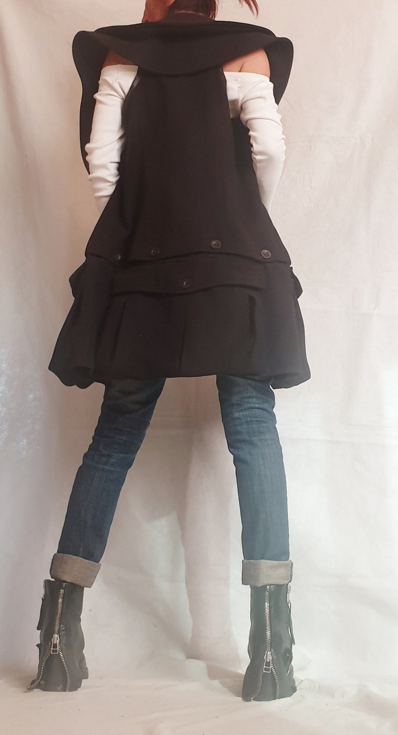 May include: A black vest with a large collar and a ruffled hem. The vest is worn over a white shirt and blue jeans. The person is wearing black boots with zippers on the sides.