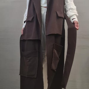 May include: A long, sleeveless dark brown vest featuring a notched collar, two chest pockets with flaps, and two large lower patch pockets. The vest includes a matching fabric tie belt at the waist and has a straight, open-front design that falls below the knees.