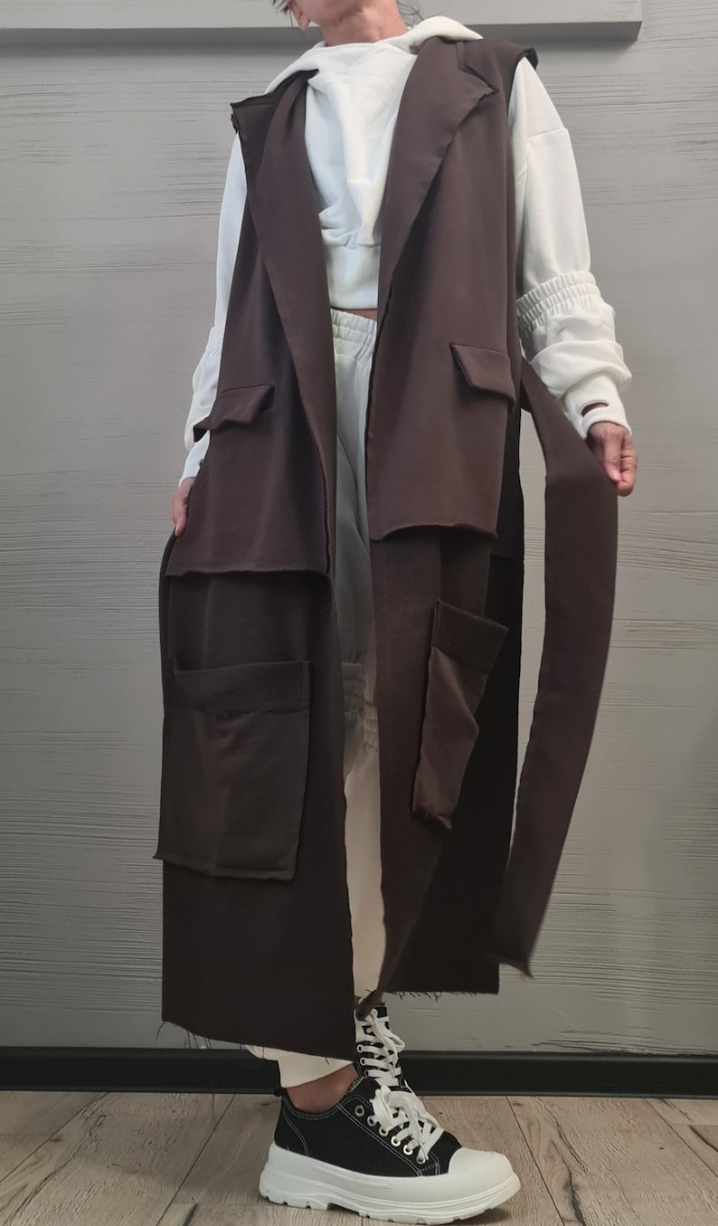May include: A long, sleeveless dark brown vest featuring a notched collar, two chest pockets with flaps, and two large lower patch pockets. The vest includes a matching fabric tie belt at the waist and has a straight, open-front design that falls below the knees.