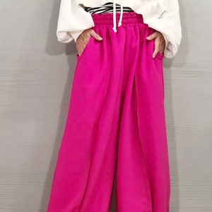 May include: A white hooded sweatshirt and bright pink wide-leg pants. The sweatshirt has a drawstring and a cropped fit. The pants have a high waist and pockets. A black and white striped shirt is visible underneath the sweatshirt.