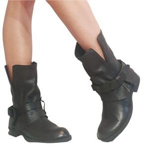 May include: Black leather ankle boots with buckle and strap detailing. The boots have a low heel and a folded cuff design. The leather appears textured. The boots are shown against a white background.
