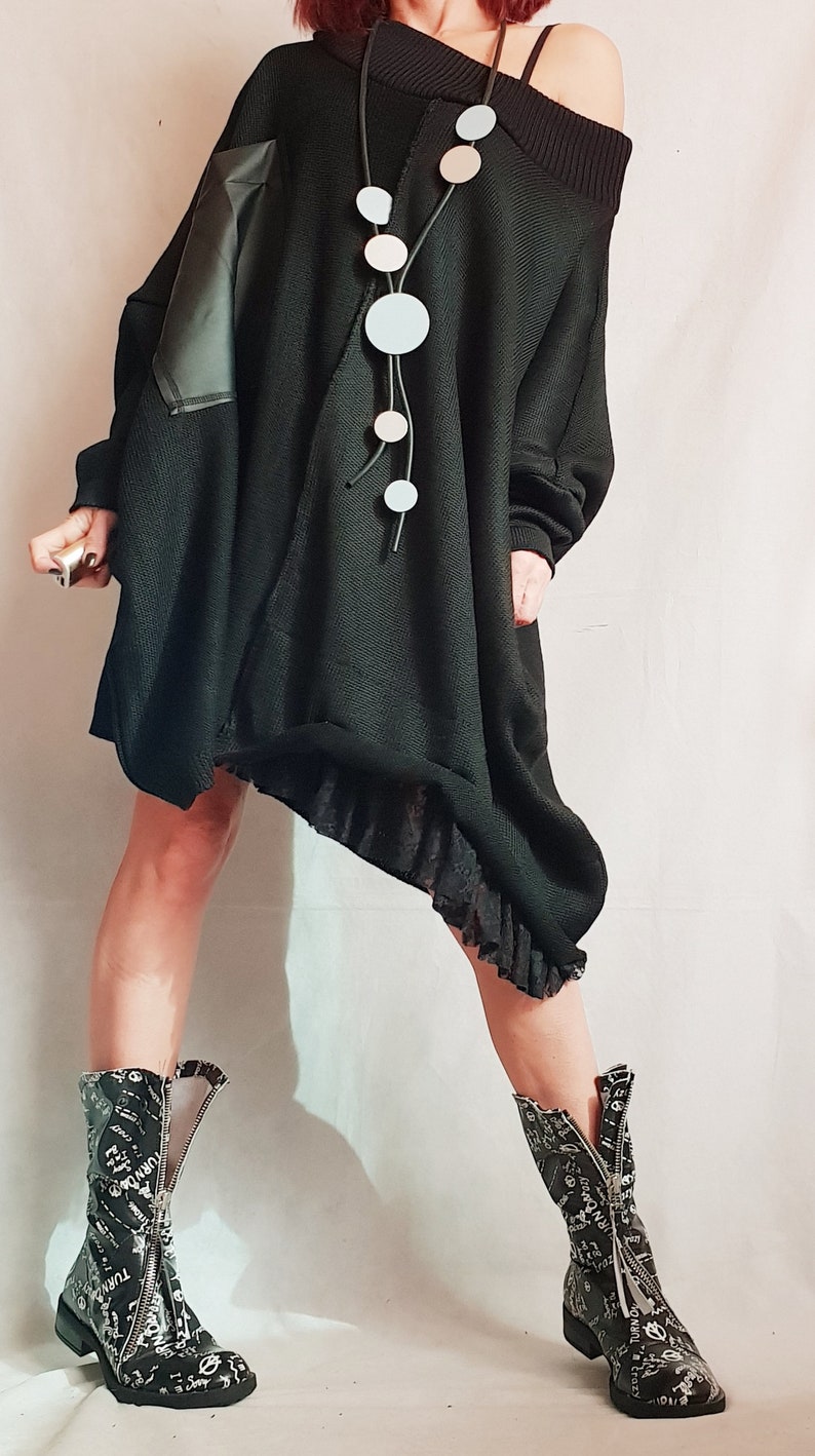 May include: An oversized black knit tunic with an asymmetrical hem, a ribbed off-the-shoulder neckline, and a black faux leather patch on the right sleeve. It is paired with a long necklace featuring circular pendants and black patterned boots.