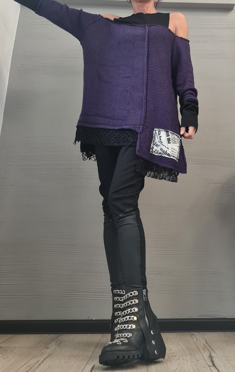 May include: A dark purple knit sweater with an asymmetrical hem, a black underlayer with lace trim, and a white patch featuring black printed text. The outfit includes black trousers and black boots with silver chain details and side zippers.