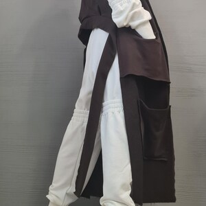 May include: A long dark brown sleeveless vest with large patch pockets worn over a white hooded sweatshirt and matching white jogger pants with gathered elastic cuffs, paired with black and white low-top sneakers with thick white rubber soles.