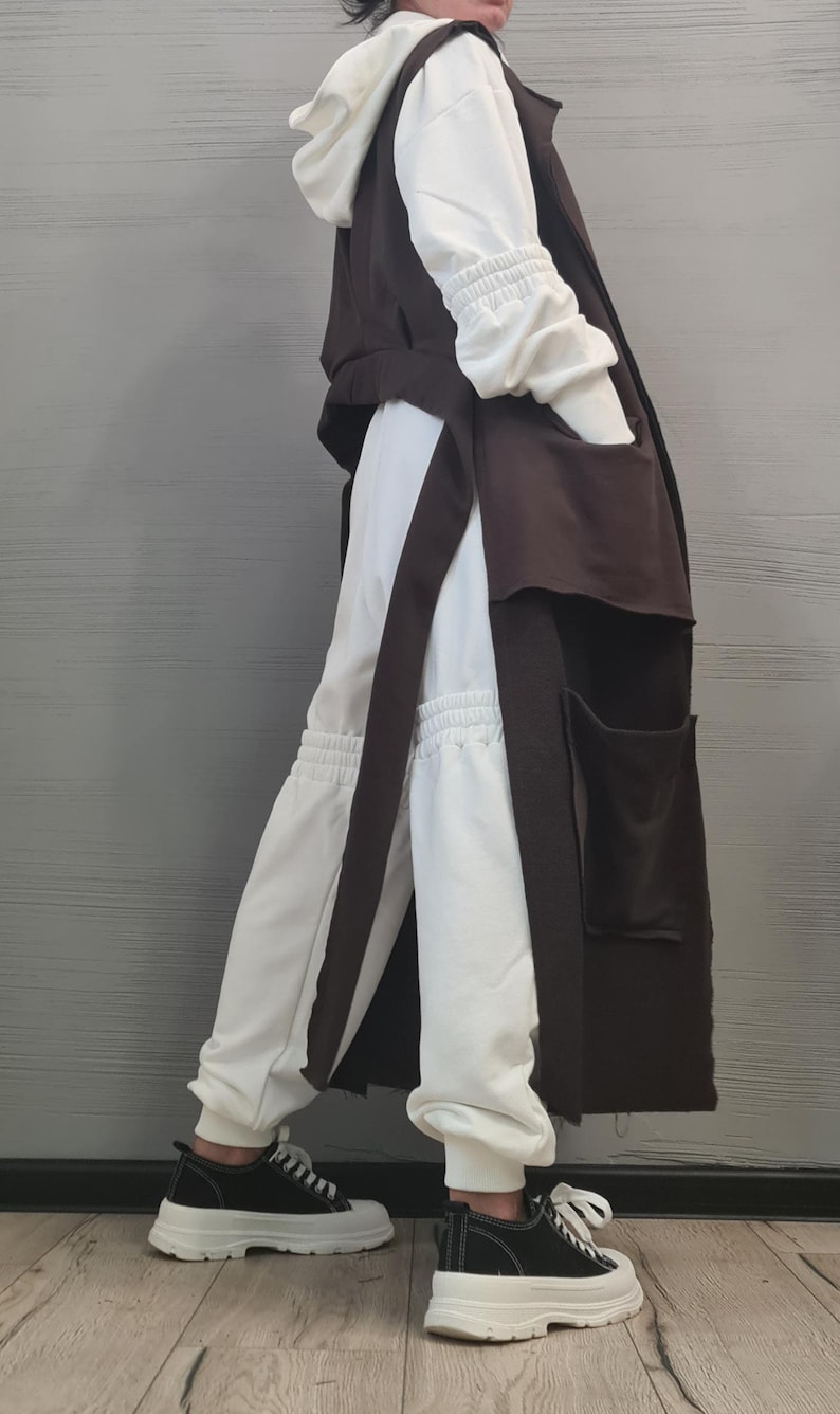 May include: A long dark brown sleeveless vest with large patch pockets worn over a white hooded sweatshirt and matching white jogger pants with gathered elastic cuffs, paired with black and white low-top sneakers with thick white rubber soles.