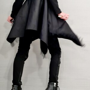 May include: A person wearing a black outfit, including a long-sleeved top with a flared skirt and black pants. The outfit is paired with black leather ankle boots with zippers and chunky heels. The background is a light gray wall.