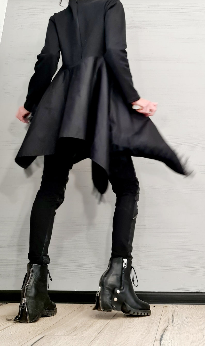 May include: A person wearing a black outfit, including a long-sleeved top with a flared skirt and black pants. The outfit is paired with black leather ankle boots with zippers and chunky heels. The background is a light gray wall.