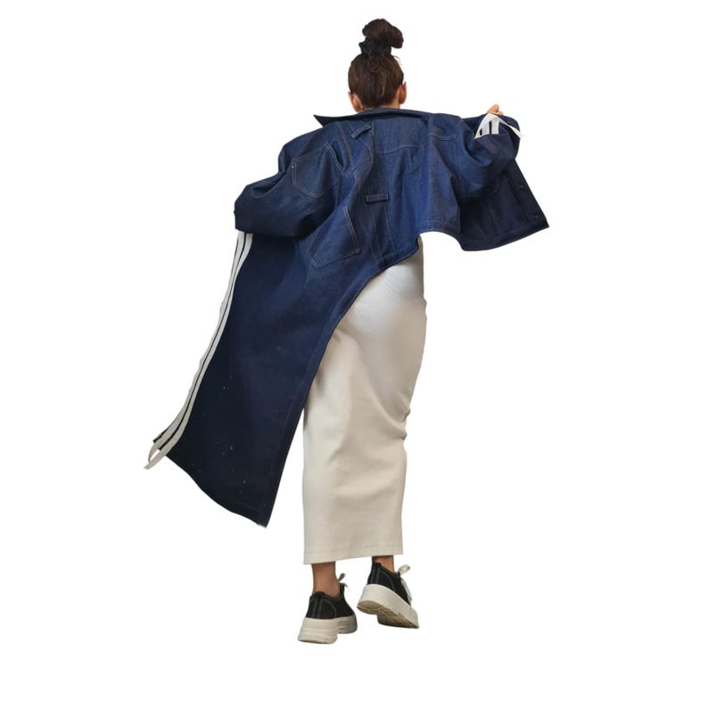 May include: A dark blue denim jacket with white stripes down the side. The jacket is long and oversized, with a classic collar and button closure. The person is wearing a white skirt and black sneakers.