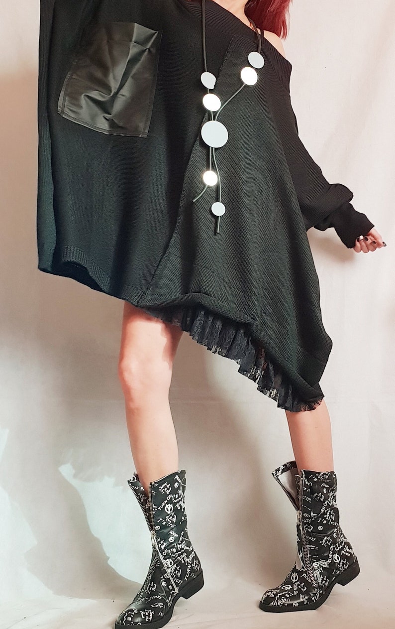 May include: An oversized black knit tunic featuring a smooth black pocket, a black lace hem, and a long necklace with circular white pendants. The outfit is paired with black mid-calf boots covered in white graphic text and symbols with front zippers.