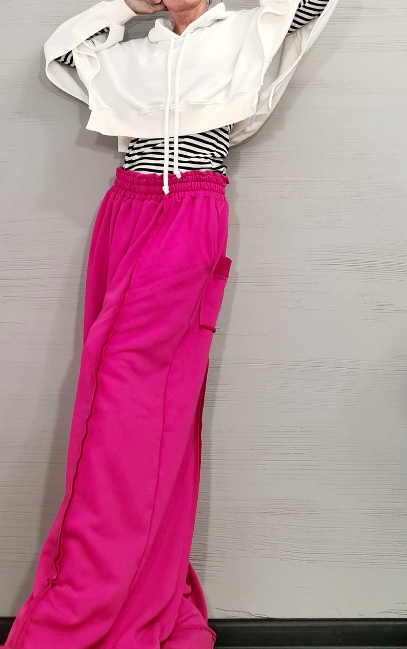 May include: A woman's outfit featuring a white hooded sweatshirt, a black and white striped long-sleeve shirt, and bright pink sweatpants. The sweatpants have a pocket on the side and a drawstring waist. The outfit is casual and comfortable.
