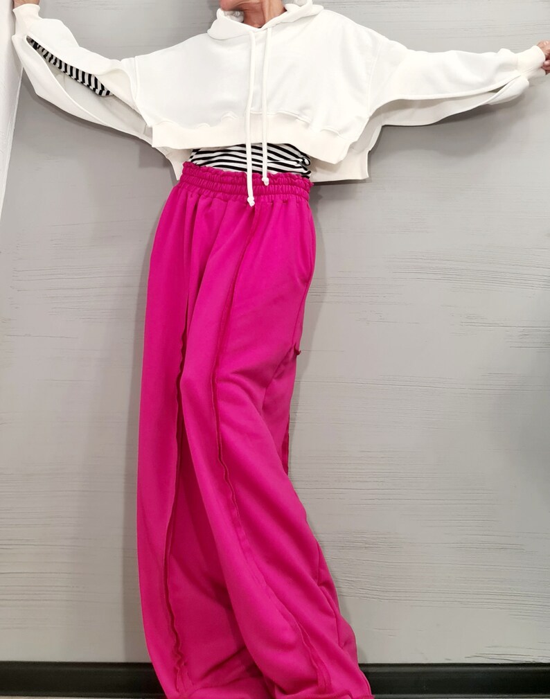 May include: A white cropped hoodie with long sleeves and a drawstring. The sleeves have a black and white striped detail. Paired with bright pink wide-leg sweatpants. A black and white striped shirt is visible underneath the hoodie.