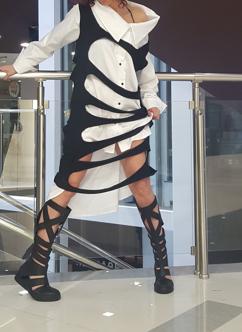 May include: A white button-down shirt is layered under a black, cut-out dress. The outfit is paired with black, open-toe, gladiator-style boots. The overall look is modern and edgy, suitable for a fashion-forward aesthetic.