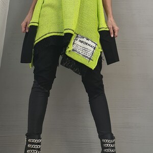 May include: A neon yellow sleeveless knit tunic with black trim and a rectangular patch featuring black text that reads "E INFORMATIO". The tunic is layered over a black top and black pants, paired with black boots featuring silver chain embellishments.