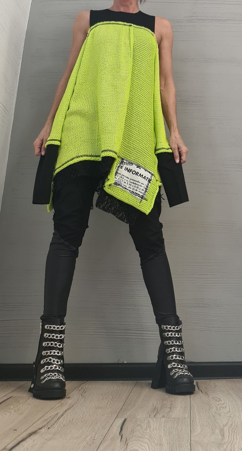 May include: A neon yellow sleeveless knit tunic with black trim and a rectangular patch featuring black text that reads "E INFORMATIO". The tunic is layered over a black top and black pants, paired with black boots featuring silver chain embellishments.