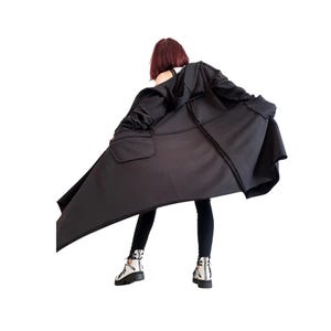 Pode incluir: Um casaco comprido preto com capuz, bainha assimétrica e um bolso grande de remendo na lateral. A peça apresenta costuras visíveis, mangas compridas e uma silhueta larga e fluida que se estende para fora do corpo.