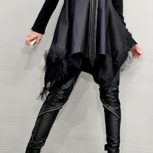 May include: Black asymmetrical tunic dress with a long-sleeved top and a flowing skirt. The outfit includes black leather-look pants with zippers, black boots, and a layered necklace. A small gold-colored object is held.