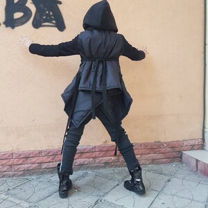 K&ouml;nnte beinhalten: Eine schwarze Kapuzenjacke mit gesteppter Westenauflage, die &uuml;ber einen Taillenbindeg&uuml;rtel und einen asymmetrischen Saum verf&uuml;gt. Dazu werden dunkelgraue, strukturierte Leggings mit wei&szlig;em Seitenstreifen und schwarze Camouflage-Stiefeletten getragen.