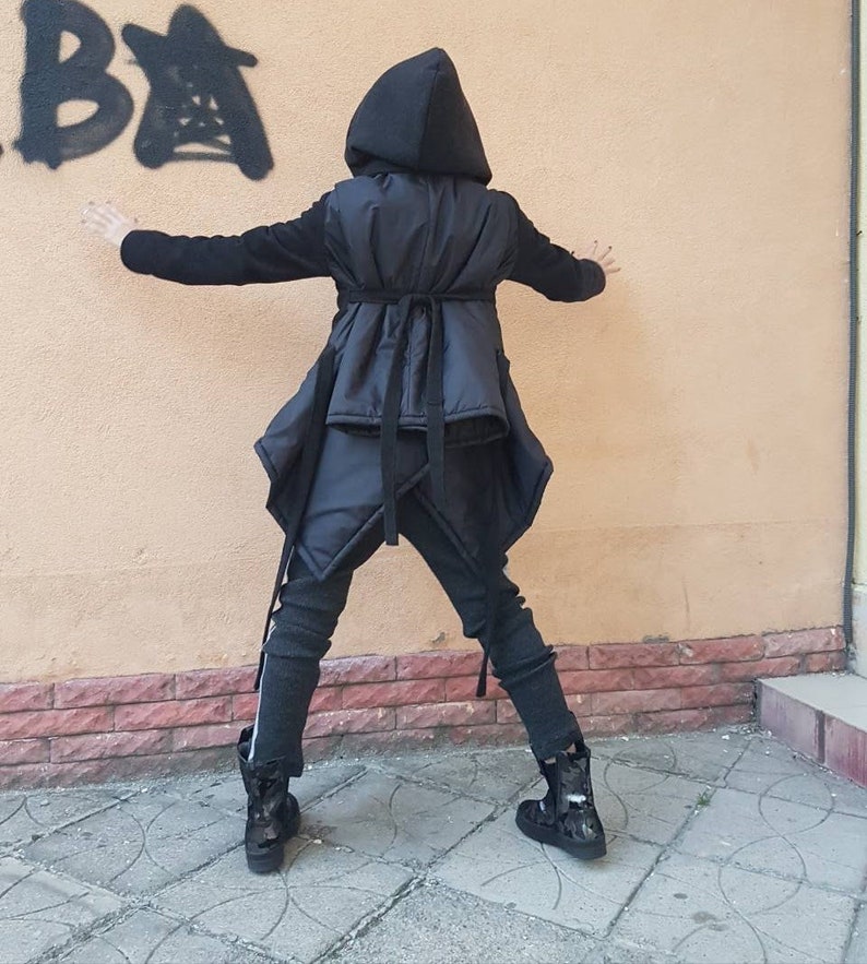 K&ouml;nnte beinhalten: Eine schwarze Kapuzenjacke mit gesteppter Westenauflage, die &uuml;ber einen Taillenbindeg&uuml;rtel und einen asymmetrischen Saum verf&uuml;gt. Dazu werden dunkelgraue, strukturierte Leggings mit wei&szlig;em Seitenstreifen und schwarze Camouflage-Stiefeletten getragen.