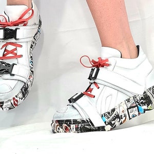 Pu&ograve; includere: Sneakers alte bianche con lacci rossi e fibbie nere. Le suole sono decorate con un collage di immagini e testi colorati. Le scarpe hanno un design moderno e audace.
