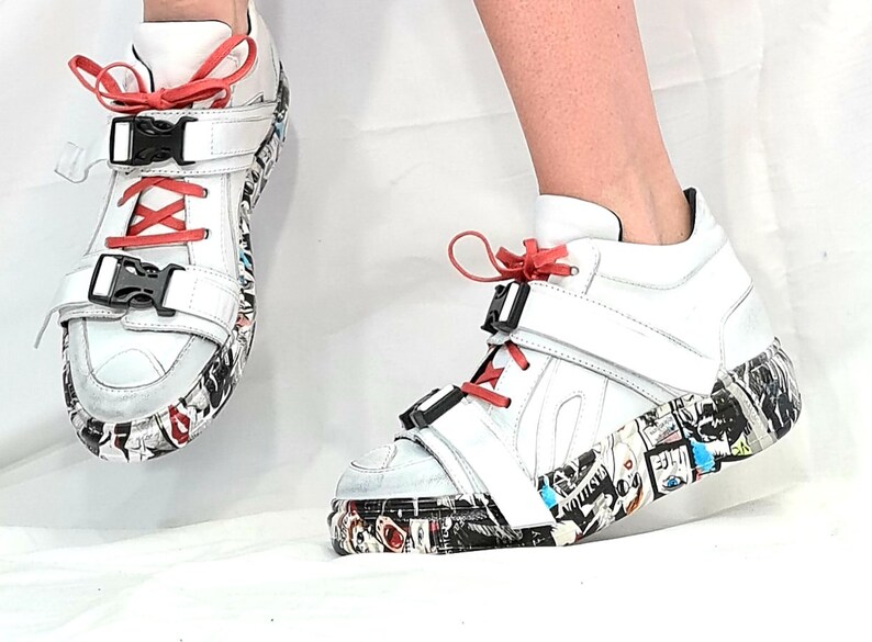 Pu&ograve; includere: Sneakers alte bianche con lacci rossi e fibbie nere. Le suole sono decorate con un collage di immagini e testi colorati. Le scarpe hanno un design moderno e audace.