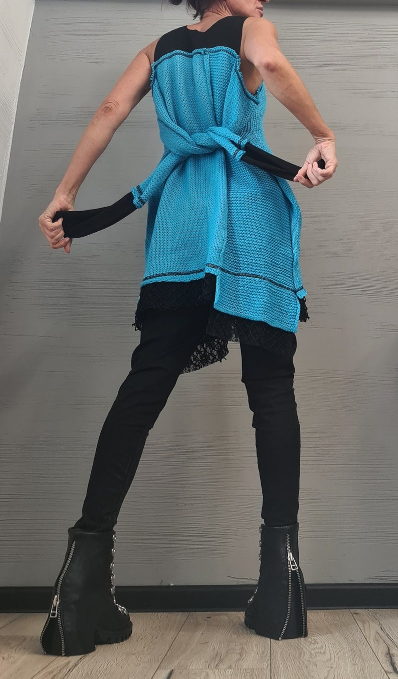 May include: A sleeveless bright blue knit tunic with a black trim and a knotted waist tie, layered over a black lace-trimmed garment and black trousers. The outfit is completed with black leather ankle boots featuring a prominent back zipper.