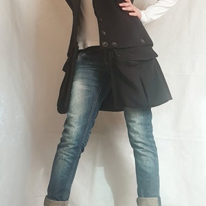 May include: A black vest with a large collar and a skirt-like bottom. The vest has two rows of buttons and is worn over a white long-sleeved shirt and blue jeans. The person is wearing black ankle boots.