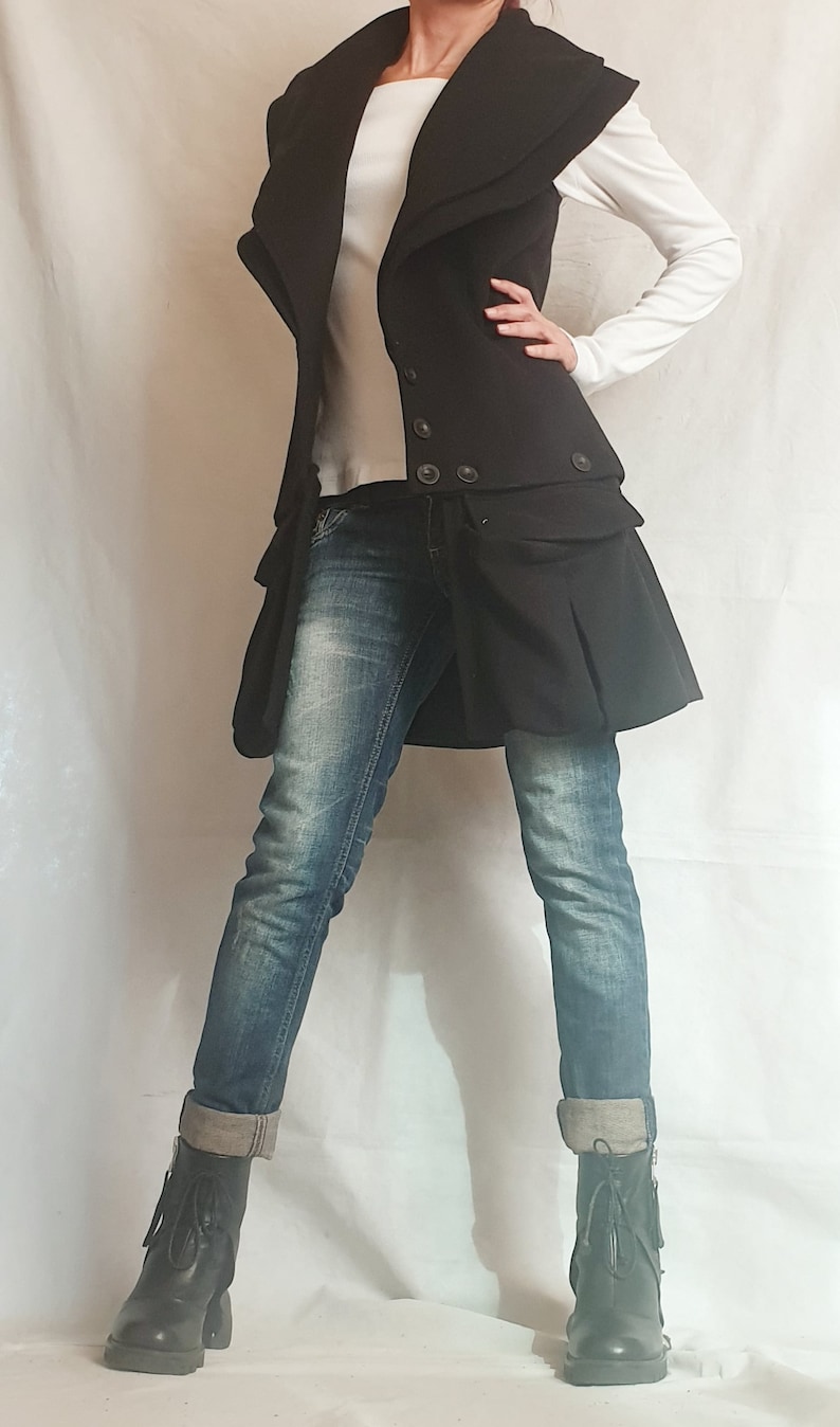 May include: A black vest with a large collar and a skirt-like bottom. The vest has two rows of buttons and is worn over a white long-sleeved shirt and blue jeans. The person is wearing black ankle boots.