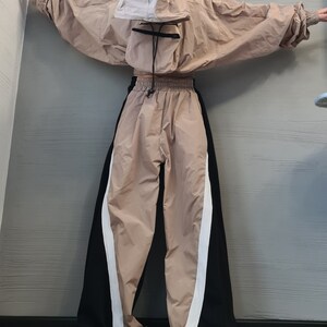 May include: A beige hooded jacket with a front pouch pocket and matching wide-leg trousers featuring black side panels and white vertical stripes. The jacket has long sleeves with elastic cuffs, and the trousers have an elastic waistband with a drawstring.