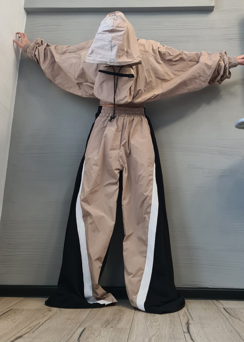 May include: A beige hooded jacket with a front pouch pocket and matching wide-leg trousers featuring black side panels and white vertical stripes. The jacket has long sleeves with elastic cuffs, and the trousers have an elastic waistband with a drawstring.