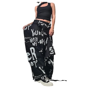 Puede incluir: Pantalones negros con estampado blanco que incluye las palabras "Young Forever". Los pantalones se combinan con un top negro y zapatillas blancas.