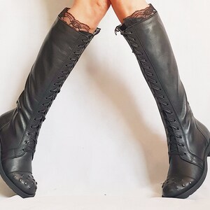 May include: Black knee-high boots with lace trim at the top. The boots have a lace-up design and small metal studs on the toe. The boots are made of a smooth, dark material.
