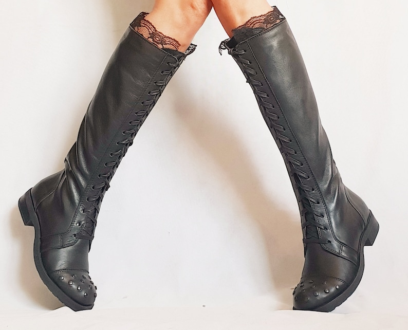 May include: Black knee-high boots with lace trim at the top. The boots have a lace-up design and small metal studs on the toe. The boots are made of a smooth, dark material.