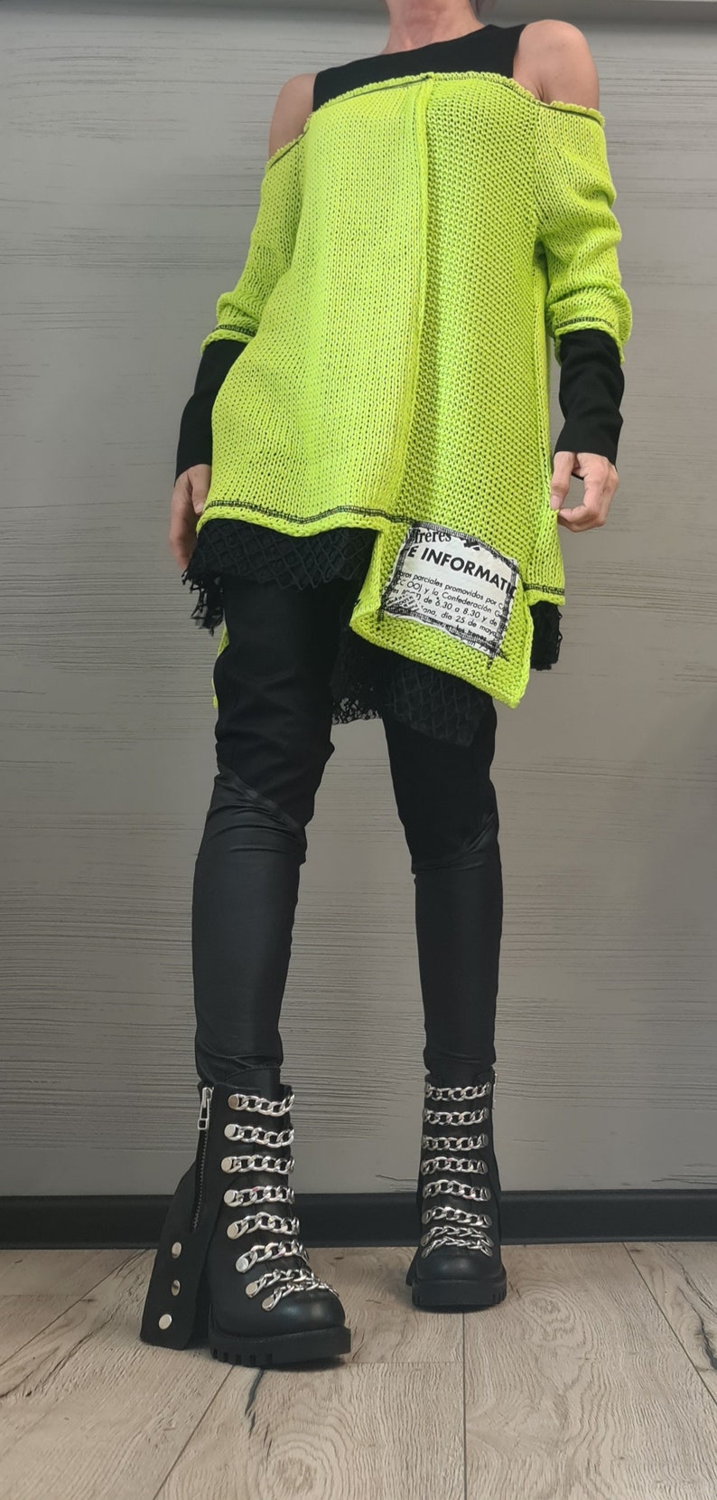 May include: A neon yellow cold-shoulder knit sweater with black long sleeves and a black lace hem, layered over black leggings and black ankle boots featuring silver chain embellishments and side zippers. A white patch with black text is attached.