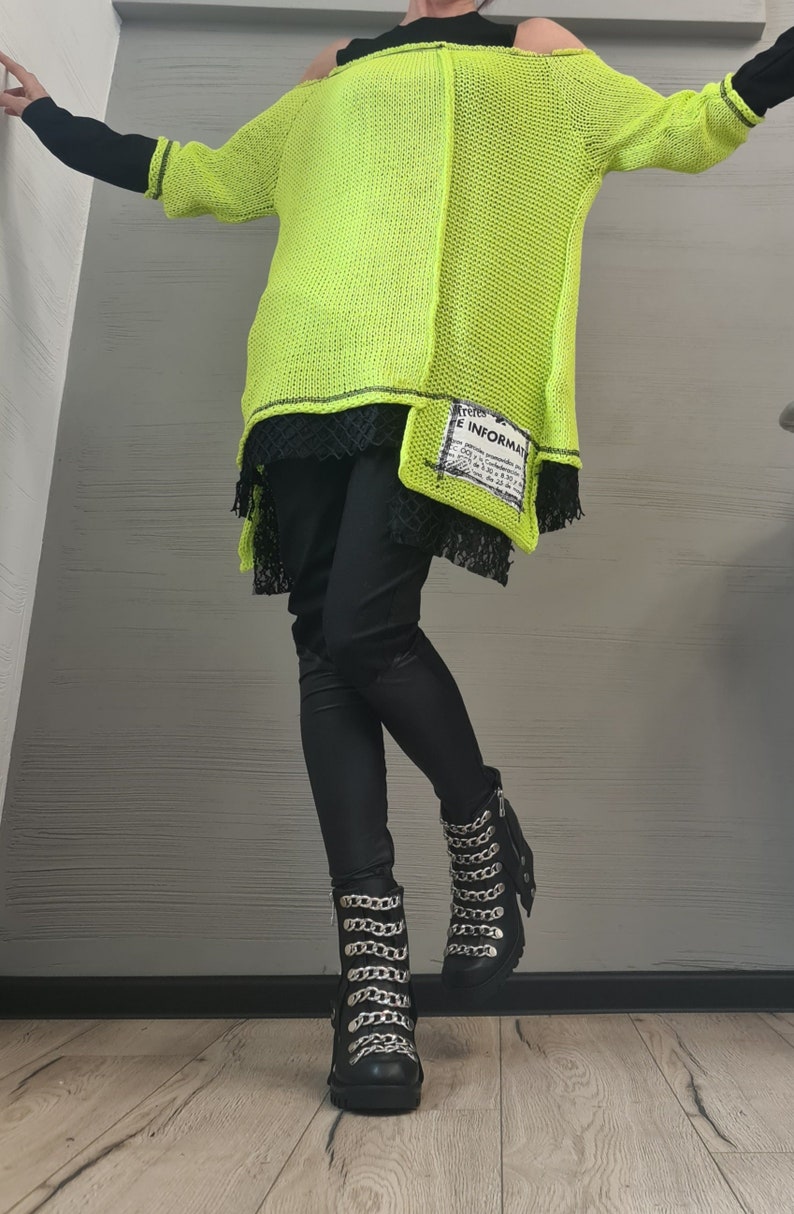 May include: A neon yellow loose-knit sweater with an off-the-shoulder neckline, layered over a black top and black lace hem. The sweater features a rectangular patch with the text "INFORMAT" and black leggings paired with black boots with silver chains.